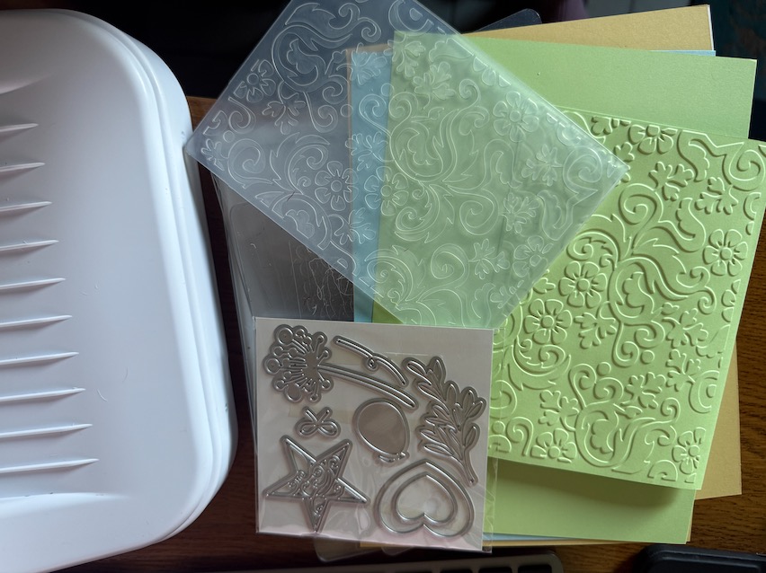 Kartenkunst: Eine Stanzmaschine mit einer geprägten Karte, einem Plastik-Prägefolder und passenden Stanzschablonen. Das Bild zeigt das kreative Handwerkszeug für die Herstellung von handgemachten Karten