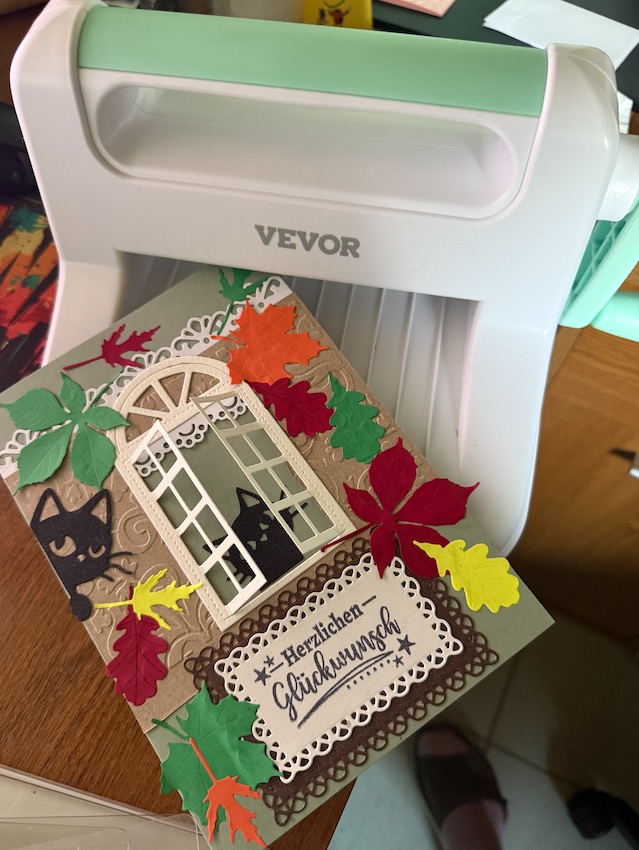 Kartenkunst: Eine fertige handgemachte Scrapbooking-Karte mit einem Katzen-Fenster-Motiv und fliegenden Blättern liegt neben der Stanzmaschine, mit der sie hergestellt wurde