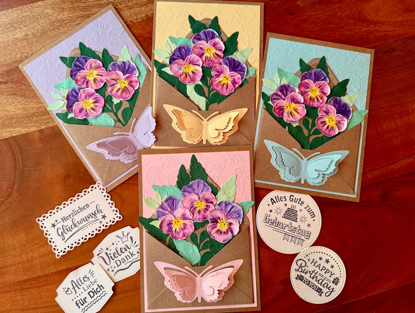 Set aus vier handgefertigten 3D-Karten in Pastellfarben. Jede Karte zeigt einen Strauß handgemalter Aquarell-Stiefmütterchen in einer Papiertasche und ist mit einem plastischen, auf den Hintergrund farblich abgestimmten Schmetterling verziert.