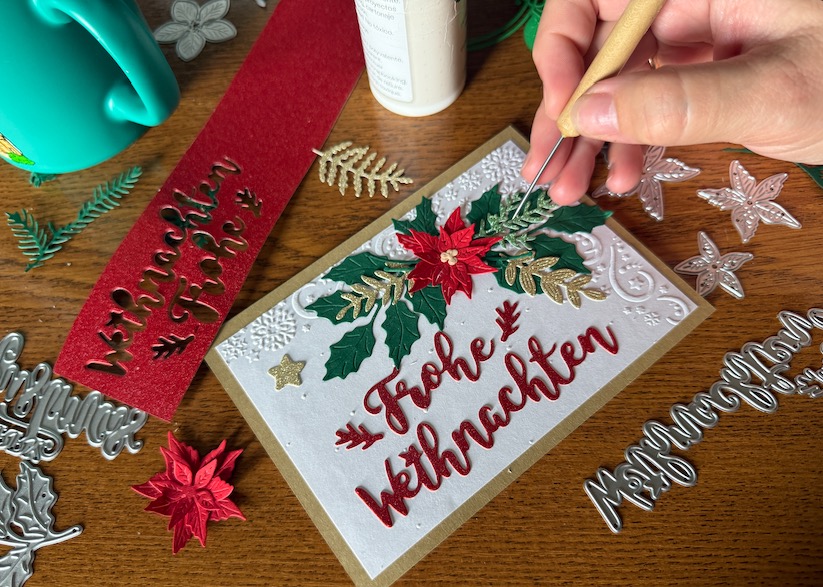 Blick hinter die Kulissen: Die liebevolle Handarbeit bei der Herstellung einer 3D-Weihnachtskarte. Eine Hand platziert mit einem Werkzeug ein goldenes Blatt auf einem Weihnachtsstern. Auf dem Holztisch sind Stanzformen und ausgestanzte Papierteile verstreut.